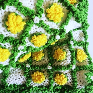 May include: A handmade crochet blanket featuring a repeating pattern of white squares, outlined in green, with yellow flower-like embellishments. The blanket has a textured appearance and a vibrant, cheerful aesthetic, perfect for home decor.
