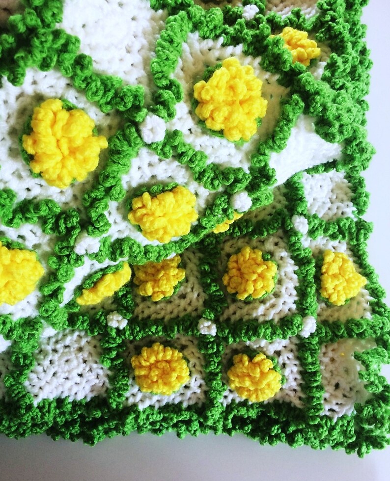 May include: A handmade crochet blanket featuring a repeating pattern of white squares, outlined in green, with yellow flower-like embellishments. The blanket has a textured appearance and a vibrant, cheerful aesthetic, perfect for home decor.