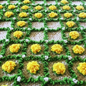 May include: A handmade crochet blanket featuring a repeating pattern of yellow pom-poms, green vines, and white accents on a cream-colored background. The blanket's design creates a textured, three-dimensional effect, perfect for home decor.
