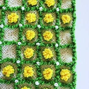 May include: Hand-knitted blanket featuring a repeating pattern of yellow flower-like clusters within green and white squares. The blanket has a textured, handmade appearance, with a bright and cheerful color palette. The design evokes a floral theme.