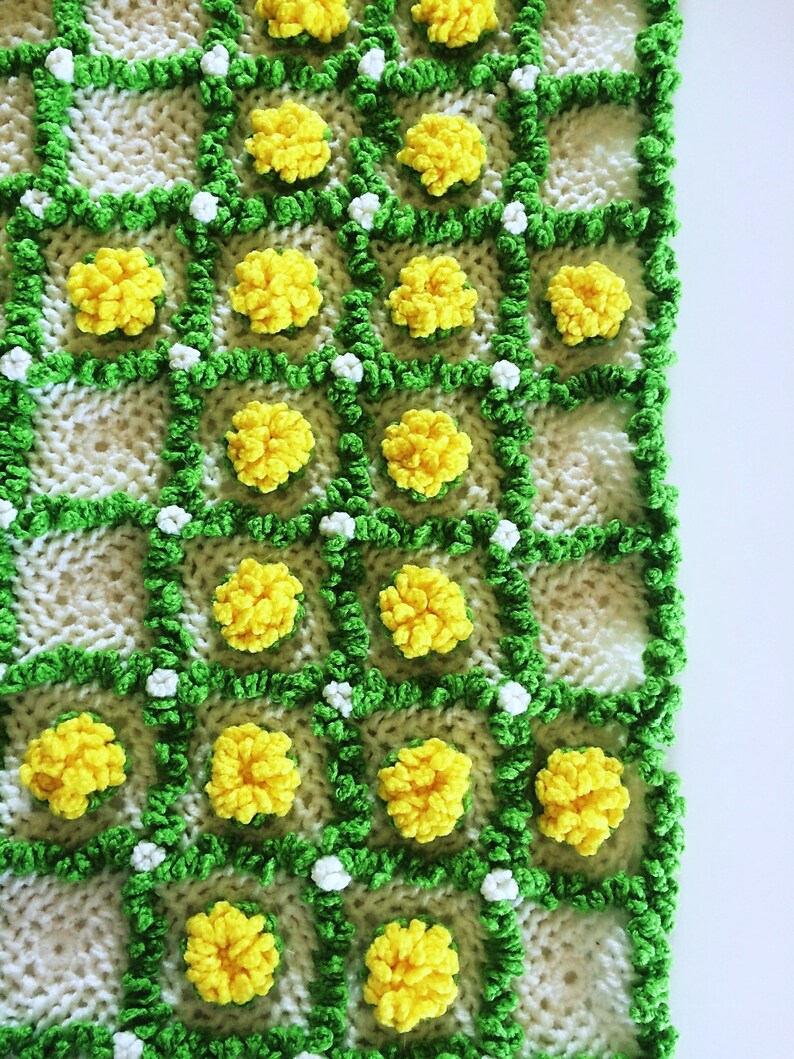 May include: Hand-knitted blanket featuring a repeating pattern of yellow flower-like clusters within green and white squares. The blanket has a textured, handmade appearance, with a bright and cheerful color palette. The design evokes a floral theme.