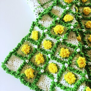 May include: A handmade crochet blanket featuring a white base with a green grid pattern. Yellow pom-pom flowers are evenly spaced throughout the design. The blanket's edge is trimmed with green yarn.