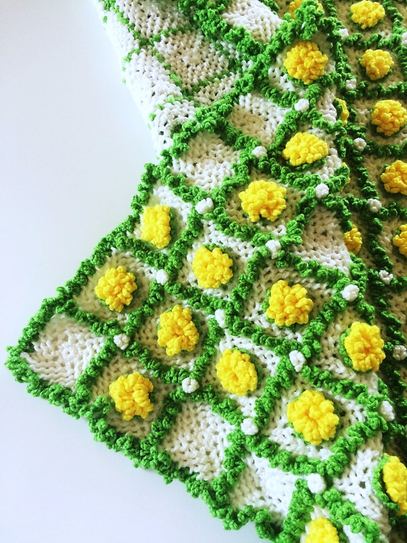 May include: A handmade crochet blanket featuring a white base with a green grid pattern. Yellow pom-pom flowers are evenly spaced throughout the design. The blanket's edge is trimmed with green yarn.