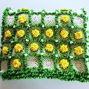 May include: A handmade, square-shaped, white knitted blanket with a repeating pattern of yellow pom-poms and green trim. The blanket has a grid-like design with the yellow pom-poms centered in each square. White and green details are also present.