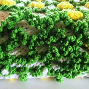 May include: Close-up of a handmade crochet blanket with a floral design. The blanket features yellow, white, and green yarn, creating a textured, three-dimensional effect. The green yarn forms a textured border, while yellow and white yarn create the floral pattern.