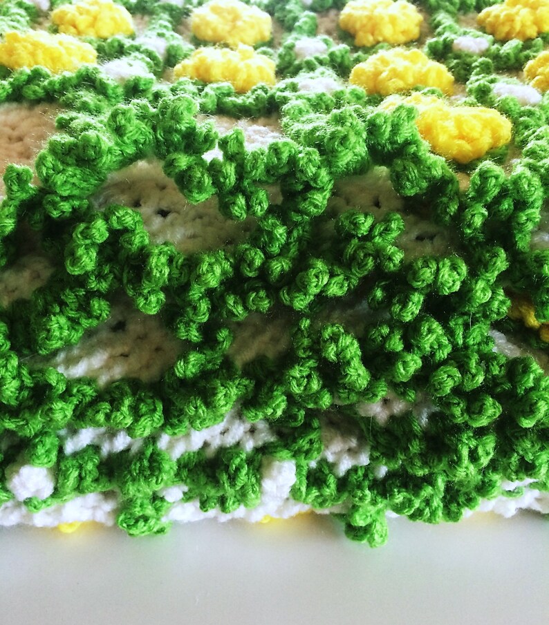 May include: Close-up of a handmade crochet blanket with a floral design. The blanket features yellow, white, and green yarn, creating a textured, three-dimensional effect. The green yarn forms a textured border, while yellow and white yarn create the floral pattern.
