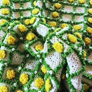 May include: Handmade crochet blanket featuring a repeating pattern of white squares with yellow flower-like accents. The squares are outlined with green and white trim, creating a textured, cozy appearance. The blanket is folded, showcasing its intricate design.