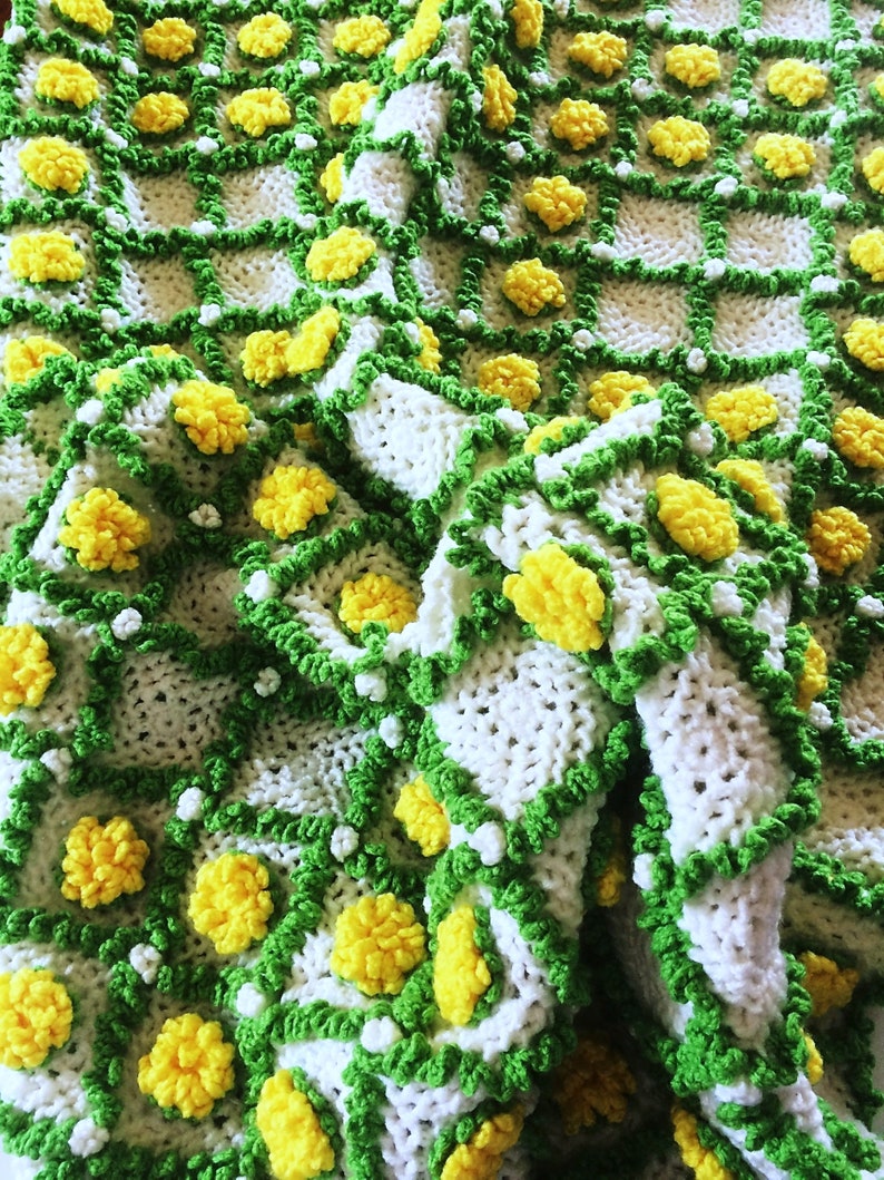 May include: Handmade crochet blanket featuring a repeating pattern of white squares with yellow flower-like accents. The squares are outlined with green and white trim, creating a textured, cozy appearance. The blanket is folded, showcasing its intricate design.