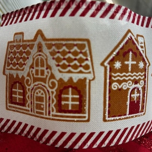 May include: A white fabric ribbon featuring a repeating pattern of brown gingerbread houses with red and white icing details. The edges of the ribbon are decorated with a red and white diagonal striped border, and the houses have glittery accents.