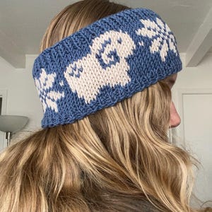 May include: A blue knitted headband with a white pattern of a sheep and snowflakes. The headband is made of a thick yarn and is worn on the head. The design is a repeating pattern across the headband.