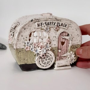 May include: A ceramic campervan ornament with the text "MY HAPPY PLACE". The campervan features a pink door with a heart, a window, and a wheel. It is adorned with pink, blue, and white flowers.