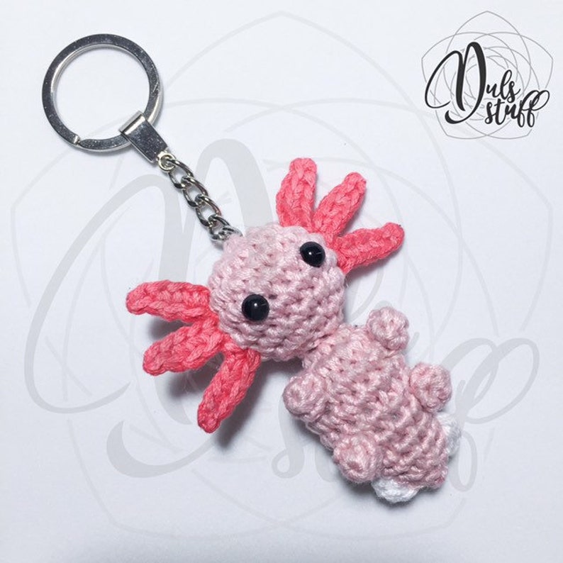 May include: A pink and red crocheted axolotl keychain with black eyes. The axolotl is attached to a silver key ring.