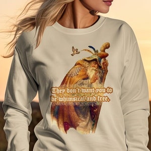 Peut inclure: Un sweat-shirt beige avec une illustration fantaisiste d'un homme barbu portant un chapeau à plumes. Le texte "They don't want you to be whimsical and free." est imprimé sur le sweat-shirt.