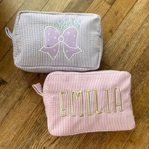 May include: Two seersucker cosmetic bags. One is gray and white striped with the name "WALDA" embroidered above a purple bow. The other is pink and white striped with the name "EMILIA" embroidered on it.