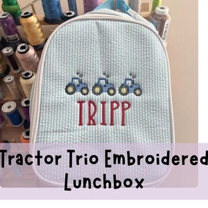 May include: A light blue and white striped lunchbox with three embroidered blue tractors and the name "TRIPP" in red. The lunchbox has a white trim and a blue handle. The text "Tractor Trio Embroidered Lunchbox" is at the bottom.