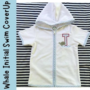 May include: White hooded swim cover-up with a blue and white striped trim and zipper. The cover-up features an embroidered letter "T" in maroon and a whale design. The text "Whale Initial Swim CoverUp" is on the left side.