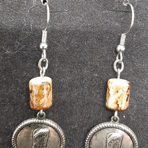 May include: A pair of silver-toned earrings featuring a cowboy boot design. Each earring has a small, cylindrical bead with brown and cream marbling, connected to a circular pendant with a boot image. The earrings hang from a silver hook.
