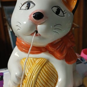 May include: A white ceramic cat figurine with black and orange accents, holding a yellow yarn ball and wearing an orange scarf. The cat has a black nose and is pulling a white string from its mouth.