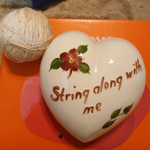 May include: A cream-colored heart-shaped ceramic piece with the words "String along with me" in brown script. It features a painted burgundy flower with green leaves and a ball of twine.