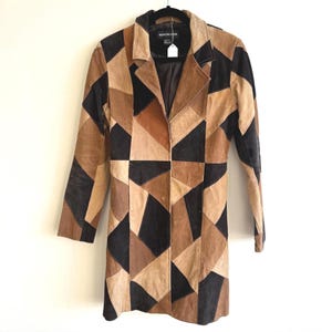May include: A long, patchwork suede coat in shades of brown and black hangs against a white background. The coat features a classic blazer style with a notched collar and long sleeves. The geometric pattern creates a unique, vintage aesthetic.