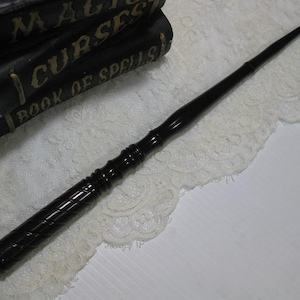 May include: A long, slender, black wooden wand with a smooth, tapered tip and a textured, spiraled handle featuring a rounded pommel at the base. The wand is positioned diagonally across the frame, showcasing its polished finish and detailed craftsmanship.