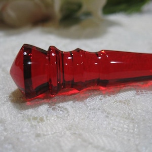 Könnte beinhalten: Ein durchsichtiger roter Glasstopfen mit einem facettierten, spitz zulaufenden Ende und drei horizontalen Rillen entlang des zylindrischen Halses. Das Glas hat eine glatte, polierte Oberfläche, die das Licht reflektiert und tiefe karmesinrote Akzente setzt.