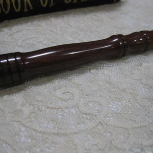 May include: A dark brown wooden wand with a faceted tip and a smooth, polished finish. The wand has a series of carved rings near the tip and a rounded handle. The wand is lying on a white lace surface.