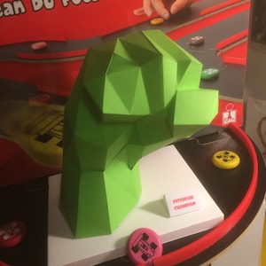 May include: A green, geometric head-shaped structure, part of a board game. The game features a red track, colourful game pieces, and the text "PITCHCAR CHAMPION".