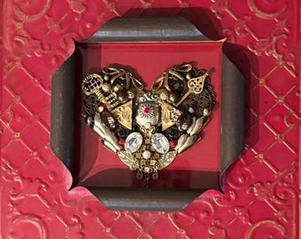 May include: A heart-shaped piece of art made from various metal and glass pieces, including keys, beads, and rhinestones. The heart is framed in a dark brown frame and set against a red background.