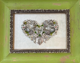 May include: A heart-shaped assemblage of vintage jewelry and embellishments, framed in a distressed green wooden frame. The heart features silver-toned metal pieces, green and clear beads, and small floral accents, creating a unique piece of art.