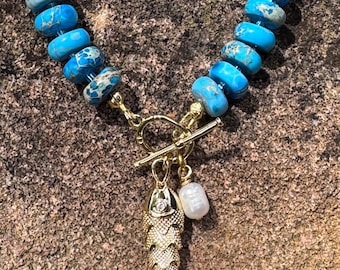 May include: A necklace featuring round, blue and teal marbled stone beads strung together. The necklace includes a gold-toned toggle clasp with a gold-toned fish pendant and a single white, irregular-shaped pearl charm hanging from the center.