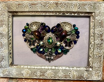 May include: A framed heart-shaped artwork made of various colorful beads, gems, and metal accents. The artwork is set against a light purple background and framed in a distressed white frame.