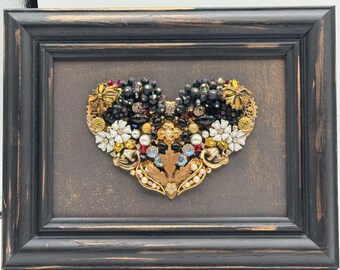 May include: Framed heart-shaped artwork composed of vintage jewelry pieces. The heart features black beads, gold-tone floral accents, white flowers, and colorful gems. The artwork is set against a brown background within a black wooden frame.