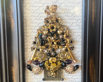 May include: A framed artwork featuring a gold and black jeweled Christmas tree made of vintage brooches, rhinestones, and other decorative elements. The background is a white paper with the words "I love you" repeated in cursive.