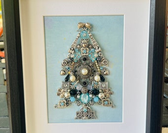 May include: A framed artwork featuring a Christmas tree made of various silver and blue jewelry pieces, including beads, buttons, and charms, on a light blue background.