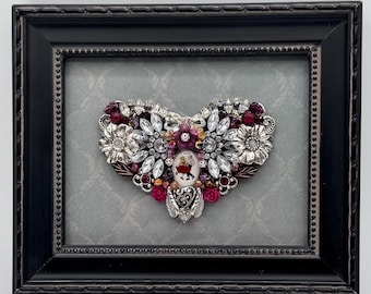 May include: A heart-shaped assemblage of vintage jewelry pieces, including clear rhinestones, silver-toned metal flowers, and deep red beads, centered around a small oval cameo depicting two figures dancing, all displayed within a black frame.