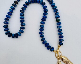 May include: A necklace featuring a long strand of round, deep blue, textured beads. The necklace is finished with a gold-toned toggle clasp that holds two gold-toned fish-shaped charms, each detailed with a small, clear, sparkling stone.