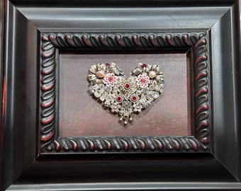 May include: A heart-shaped piece of art made from various silver and pink jewelry pieces, including rhinestones, beads, and flowers. The art is framed in a black frame with a red-brown mat.