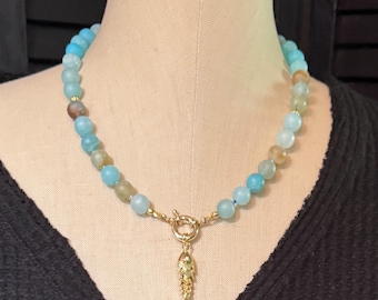 May include: A beaded necklace featuring round, matte light blue and translucent tan beads strung together. The necklace includes a gold-toned toggle clasp at the center, from which hangs a small, textured gold-toned fish-shaped pendant charm.