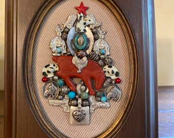 May include: A framed mixed media artwork featuring a red horse figurine surrounded by turquoise and silver beads, charms, and other decorative elements. The artwork is in a brown oval frame with a gold trim.