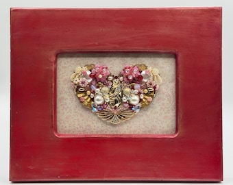 May include: A red wooden framed heart-shaped artwork. The heart is adorned with an array of pink, gold, and iridescent embellishments, including beads, pearls, and floral accents. The background features a patterned design.