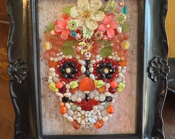 May include: A framed mosaic artwork depicting a sugar skull made of colorful beads, rhinestones, and other decorative elements. The skull is surrounded by a floral design.