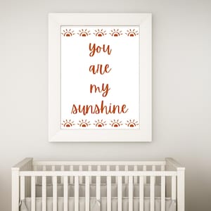 Puede incluir: Impresión enmarcada en blanco con el texto "You are my sunshine" en letras marrones. El texto está rodeado por un borde de pequeños iconos de sol marrones.