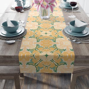 Op de afbeelding: Een rechthoekige tafelloper met een herhalend abstract patroon in tinten mosterdgeel, saliegroen en crème. De stof toont een symmetrisch, vloeiend ontwerp dat over het midden van een houten eettafel met kommen op de tafel loopt.