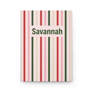 Könnte beinhalten: Ein rechteckiges Notizbuch mit einem vertikalen Streifenmuster in den Farben Rot, Dunkelgrün, Hellrosa und Creme. Der Name Savannah ist in dunkelgrünen, serifenlosen Groß- und Kleinbuchstaben in der Mitte des Einbands aufgedruckt.