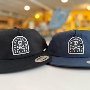 Puede incluir: Se muestran dos gorras de béisbol, una negra y otra azul marino. Cada gorra presenta un parche blanco con un diseño de calavera y huesos cruzados, y las palabras "GREEN MOUNTAIN PIRATE". Las gorras tienen una visera plana y cierre snapback.