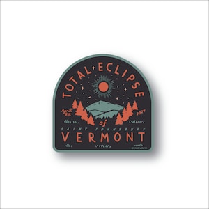 Könnte beinhalten: Ein blaugrüner und schwarzer runder Aufkleber mit dem Text "TOTAL ECLIPSE" in weißen Buchstaben. Der Aufkleber zeigt eine stilisierte Illustration einer Bergkette mit einer roten Sonne und Sternen im Nachthimmel. Der Text "April 8th, 2024" ist ebenfalls enthalten. Der Aufkleber lautet "of SAINT JOHNSBURY VERMONT".