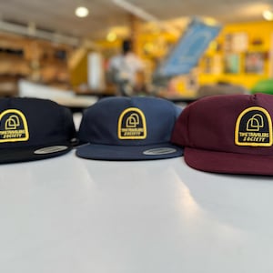 May include: Three flat-brimmed baseball caps in navy, dark blue, and burgundy. Each hat features a yellow patch with the words "TIME TRAVELERS SOCIETY" and a stylized logo. The hats are displayed on a white surface.