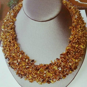 May include: A handcrafted necklace displayed on a gray mannequin. The necklace is made of small, irregularly shaped, amber-colored stones and tiny gold beads. The stones are clustered together, creating a textured, eye-catching design.
