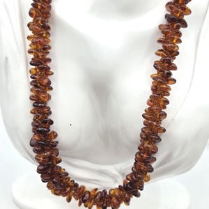 May include: A long necklace composed of numerous small, polished, translucent amber beads in varying shades of deep cognac and honey brown. The beads are strung together in a dense, multi-layered arrangement, creating a textured, organic appearance.